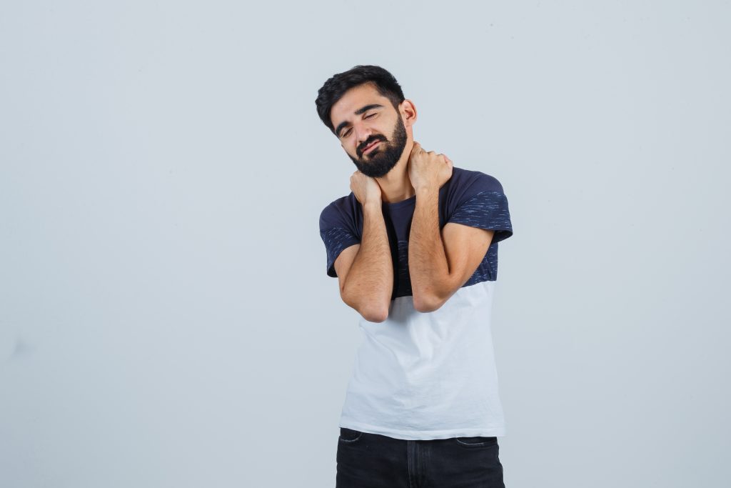 young male keeping hands on neck in t-shirt, pants and looking tired. front view.